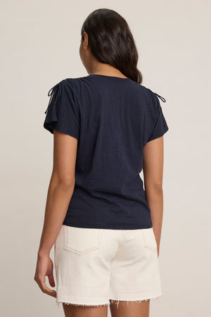 A person stands facing away, wearing the PAULA TEE by Velvet by Graham & Spencer—a navy cotton slub crew-neck with flutter sleeves—paired with light, frayed hem shorts against a plain background.