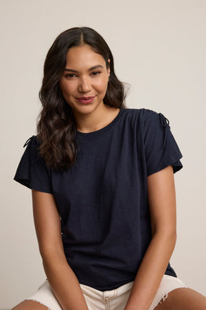 A woman with long dark wavy hair wears the PAULA TEE by Velvet by Graham & Spencer, paired with light shorts, sitting before a plain background and looking at the camera with a slight smile.