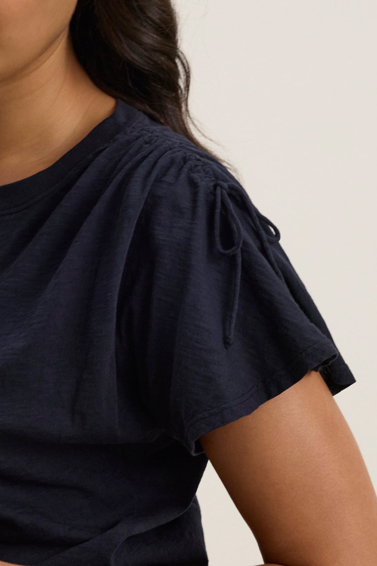 A person wears the PAULA TEE by Velvet by Graham & Spencer, a black cotton slub crew-neck style with gathered shoulders and tie detail; only the upper torso and part of the face are visible.