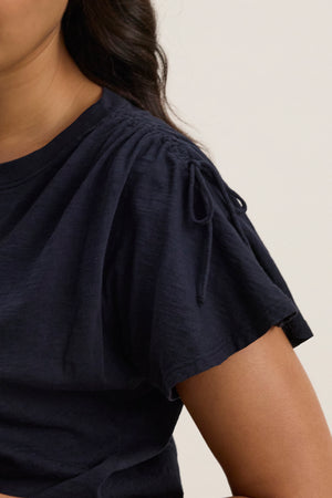 A person wears the PAULA TEE by Velvet by Graham & Spencer, a black cotton slub crew-neck style with gathered shoulders and tie detail; only the upper torso and part of the face are visible.