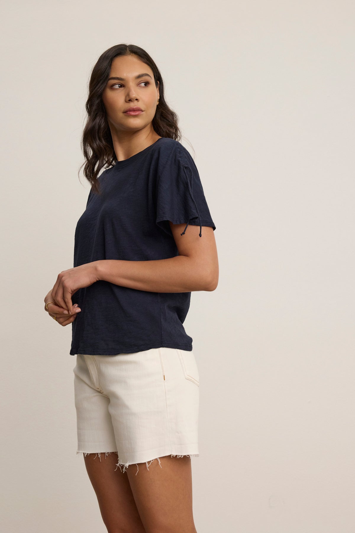 A woman stands against a plain background, wearing the PAULA TEE by Velvet by Graham & Spencer—a navy cotton slub crew-neck with short sleeves—paired with white denim shorts, looking off to the side.-39123351994561