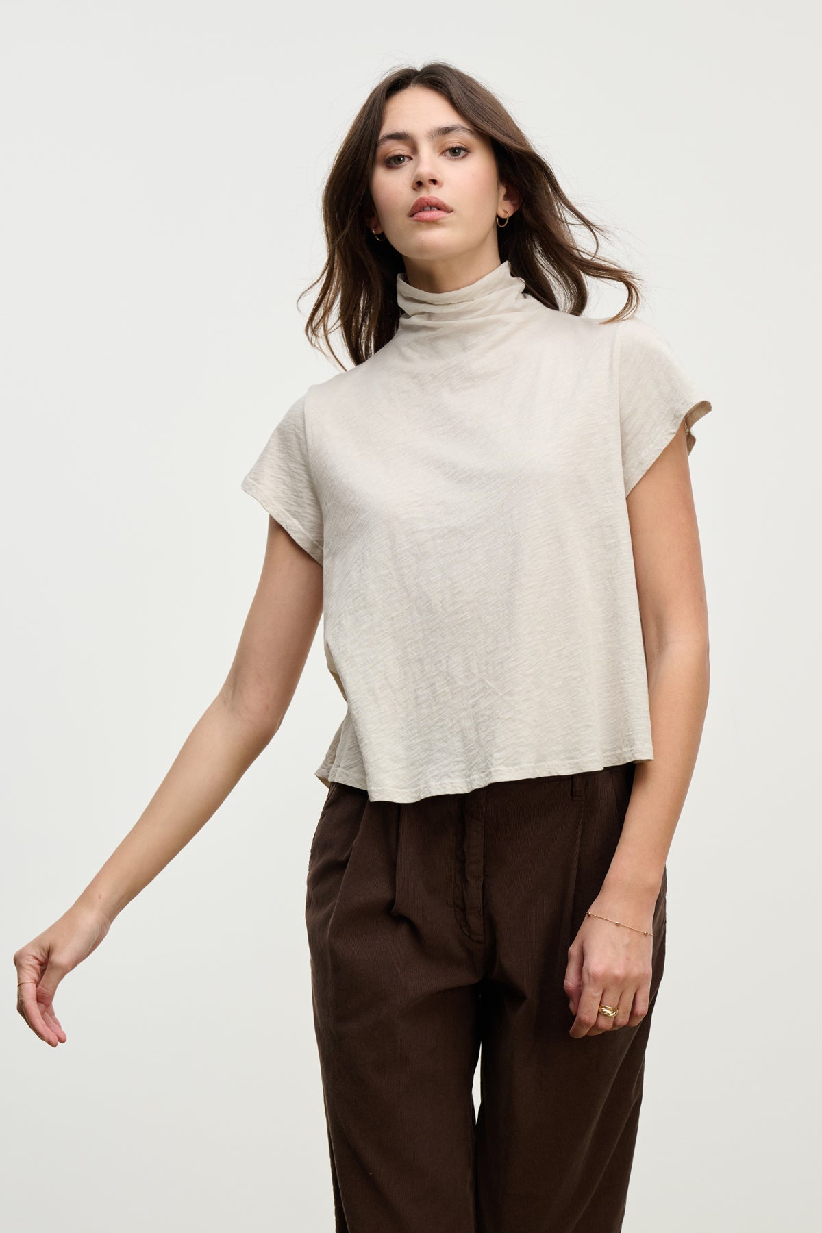 A woman wearing the SAVI TEE by Velvet by Graham & Spencer—a beige cotton slub tee with a relaxed turtleneck—and brown pants stands against a plain light background.-39800374034625