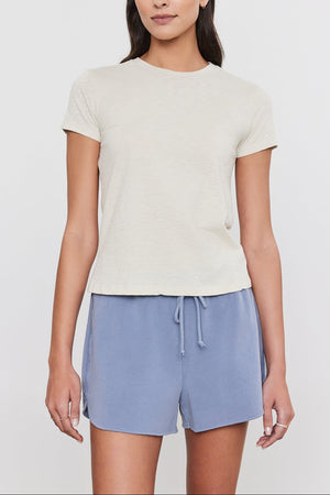 A person with long dark hair is wearing a light beige SIERRA CREW NECK TEE by Velvet by Graham & Spencer and light blue drawstring shorts, standing against a plain white background, exuding a subtle retro vibe.