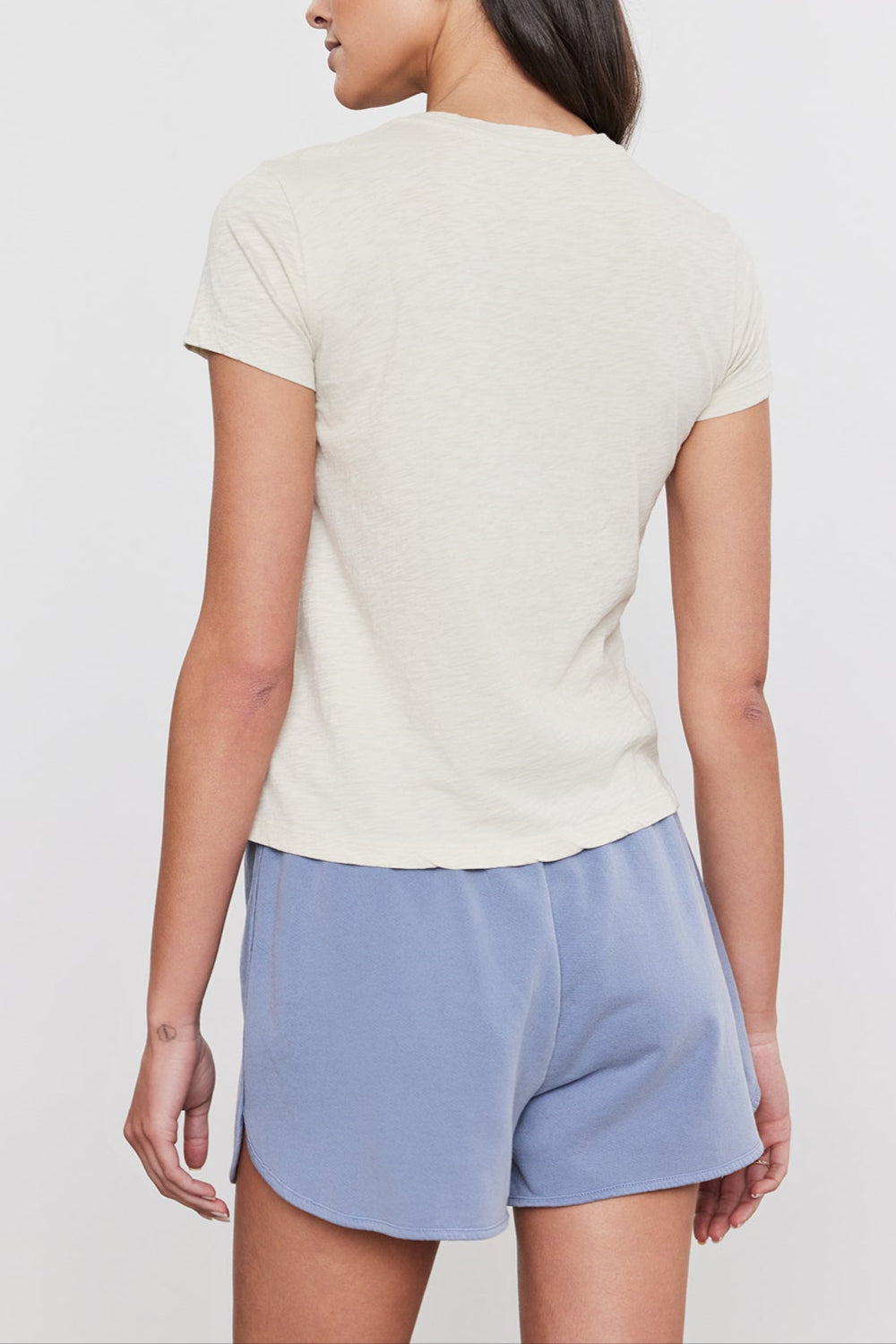 A woman with long dark hair stands with her back to the camera, wearing a short-sleeve beige SIERRA CREW NECK TEE by Velvet by Graham & Spencer and light blue shorts against a plain background, capturing a subtle retro vibe.-37452684656833