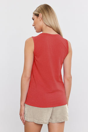 A woman with blonde hair stands with her back turned, wearing an orange TAURUS TANK TOP by Velvet by Graham & Spencer made of textured cotton slub and beige shorts.