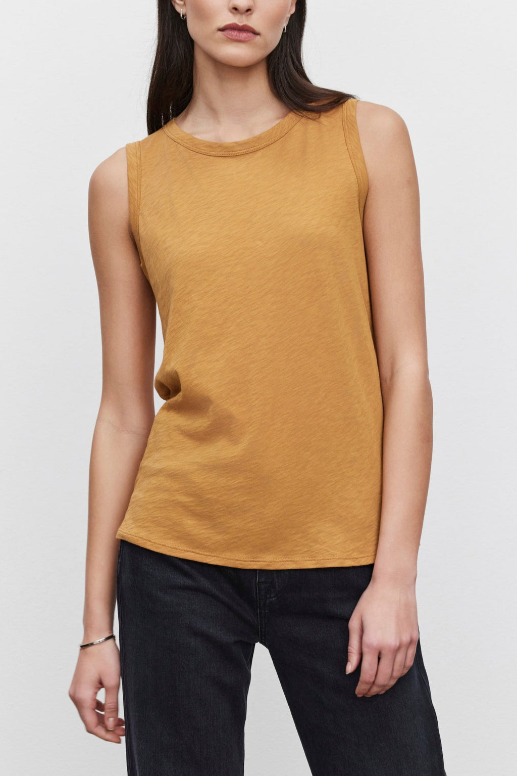 A person with long dark hair wears the TAURUS TANK TOP by Velvet by Graham & Spencer, a sleeveless, mustard-colored, crew neck top, paired with black pants while standing against a plain white background.-37629572677825