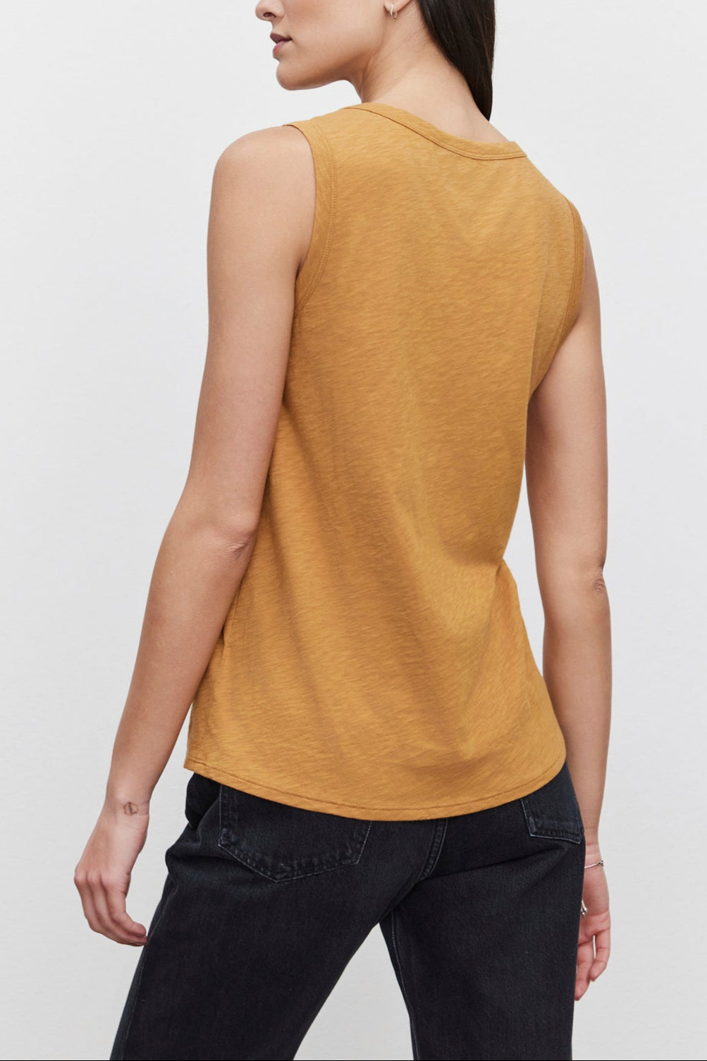 A woman with long dark hair, facing away, wearing the TAURUS TANK TOP by Velvet by Graham & Spencer, paired with dark jeans against a plain white background.-37629572743361