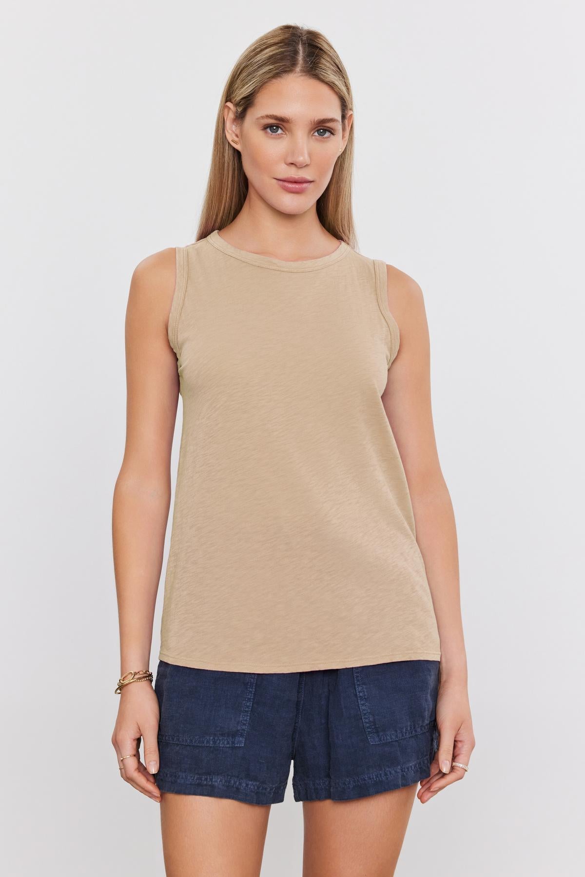 A person with long hair stands facing forward, wearing a beige sleeveless top and blue shorts against a plain white background, epitomizing the versatility of the TAURUS TANK TOP by Velvet by Graham & Spencer.-36806822527169