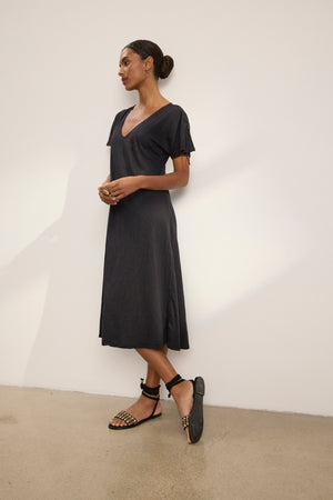 A woman stands against a plain wall, wearing the VERONICA DRESS by Velvet by Graham & Spencer— a versatile short-sleeve black midi t-shirt dress—paired with black strappy flat sandals, her hair styled in a neat bun.