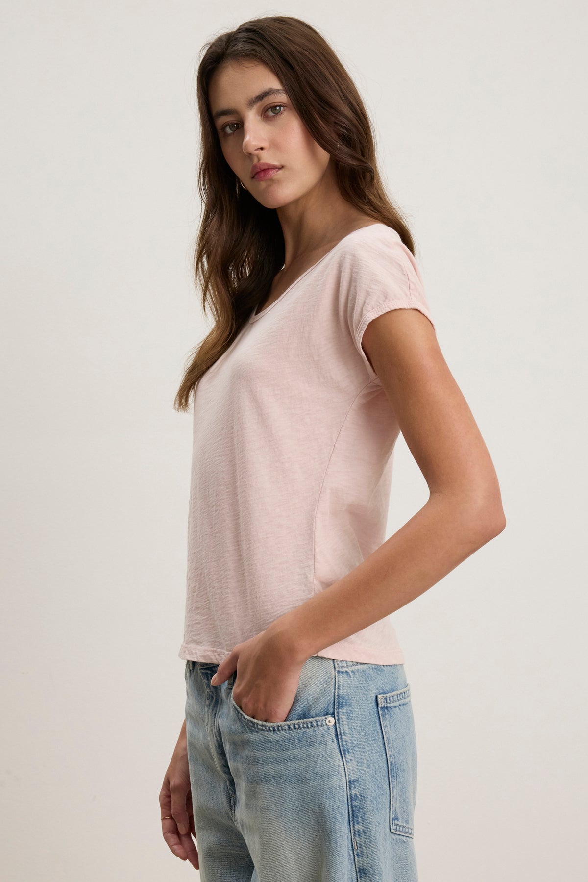 A woman stands in profile with one hand in her jeans pocket, wearing the VIVIENNE TEE by Velvet by Graham & Spencer—a light pink cotton slub tee with a scoop neckline—against a plain background.
