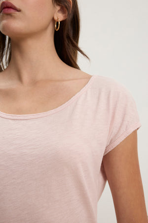 A woman wears the Velvet by Graham & Spencer VIVIENNE TEE, a light pink, short-sleeved cotton slub scoop neck top, paired with gold hoop earrings; she is shown from the shoulders up with her face partially out of frame.