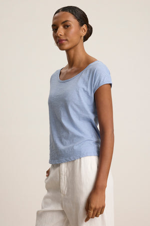 A woman poses in a relaxed stance, wearing the VIVIENNE TEE by Velvet by Graham & Spencer with a scoop neckline and white pants, set against a plain beige background.