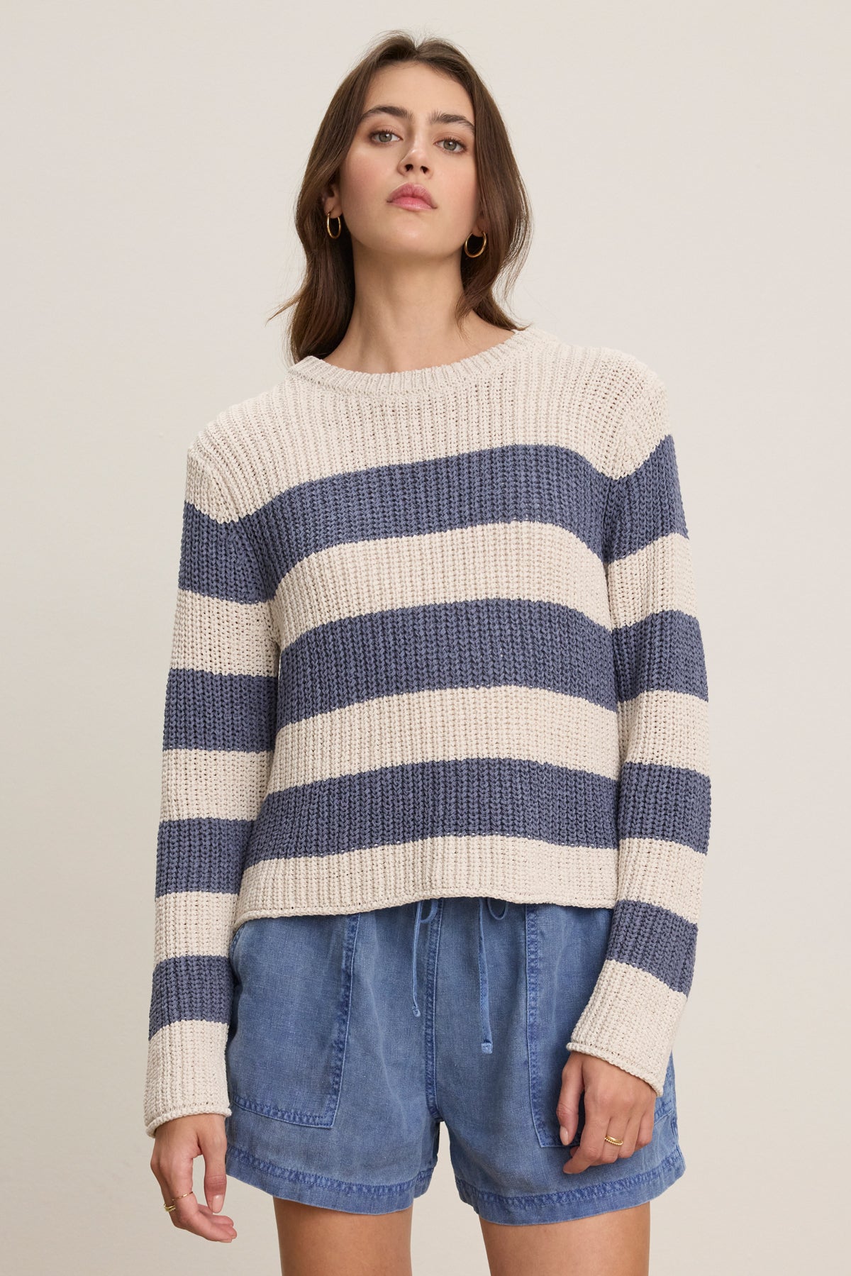 A person wears the Velvet by Graham & Spencer MAXINE SWEATER, a cream and navy striped cotton blend crewneck, paired with blue drawstring shorts, standing against a plain light background.