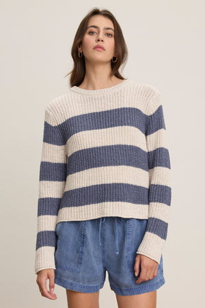 A person wears the Velvet by Graham & Spencer MAXINE SWEATER, a cream and navy striped cotton blend crewneck, paired with blue drawstring shorts, standing against a plain light background.