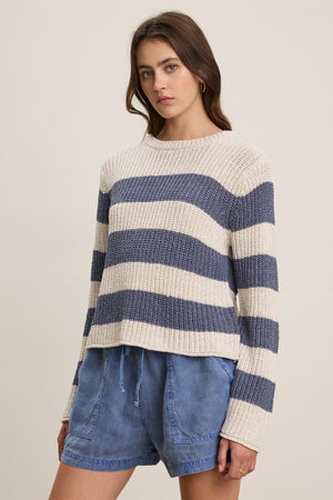 A woman wears the MAXINE SWEATER by Velvet by Graham & Spencer, a cream and blue striped cotton blend crewneck, paired with blue drawstring shorts against a plain background.