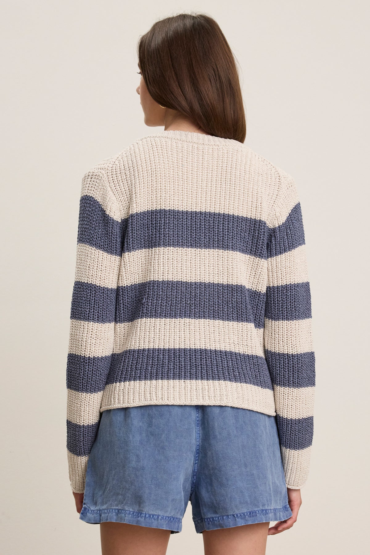 A person with straight brown hair is shown from the back wearing Velvet by Graham & Spencer’s MAXINE SWEATER—a cream and navy striped cotton blend crewneck—with blue denim shorts, against a neutral background.