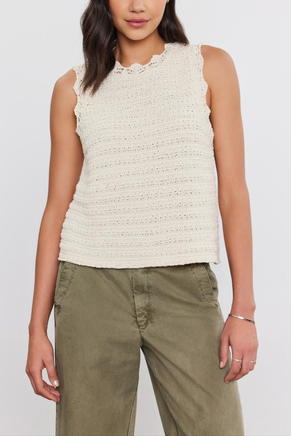 A person stands against a plain background wearing a sleeveless, cream-colored Velvet by Graham & Spencer SOPHIE SWEATER VEST with scallop details and green pants.-37054560403649