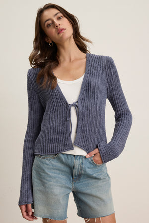 A woman wears the THALIA CARDIGAN by Velvet by Graham & Spencer, a blue long sleeve knit, layered over a white top and denim shorts against a plain background.