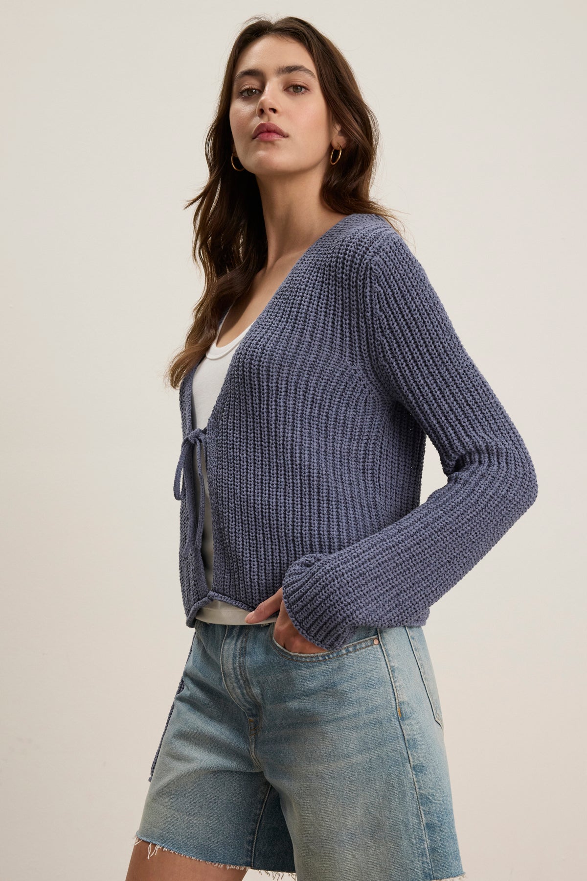A woman wears the THALIA CARDIGAN by Velvet by Graham & Spencer in blue over a white top and denim shorts, standing against a plain light background.-39111853736129