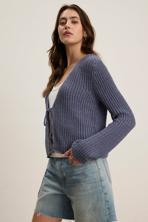 A woman wears the THALIA CARDIGAN by Velvet by Graham & Spencer in blue over a white top and denim shorts, standing against a plain light background.