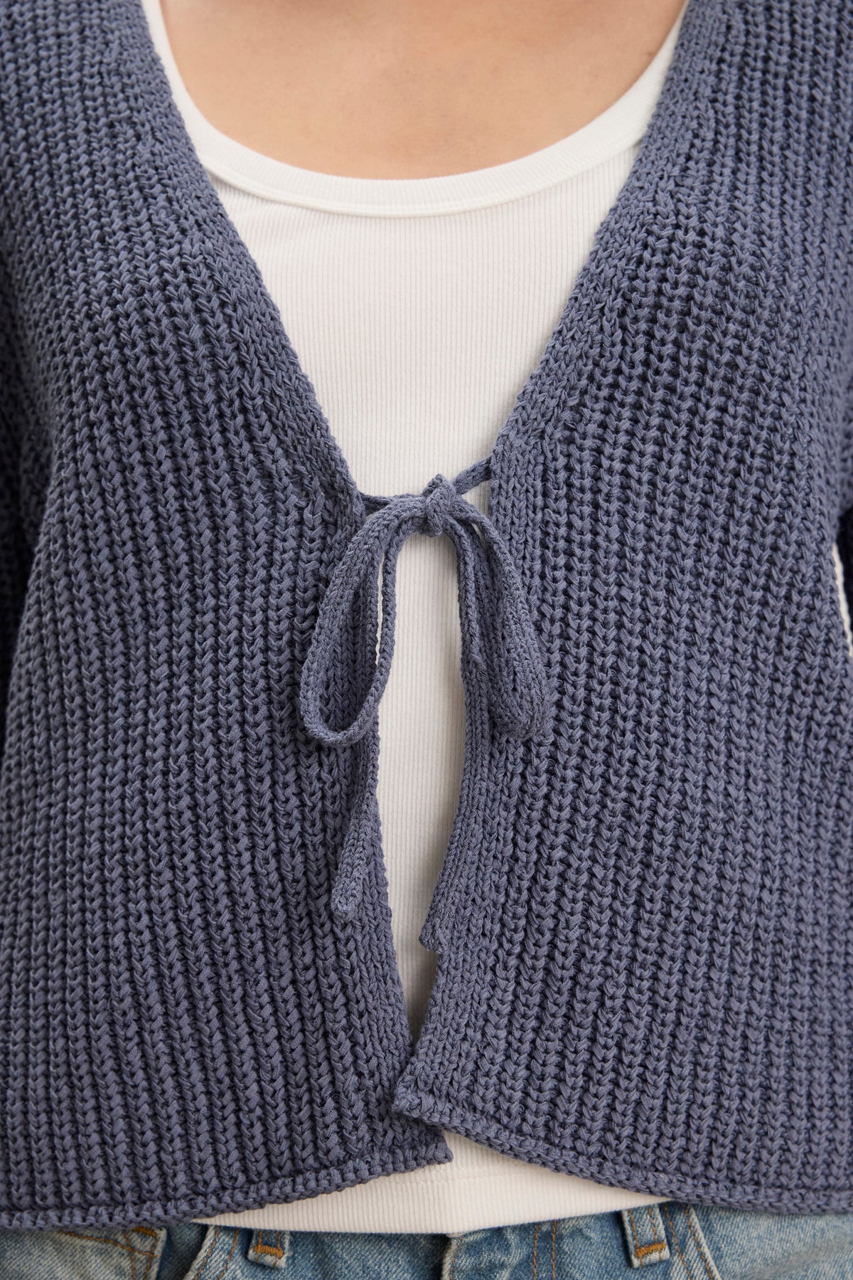 A close-up of a person wearing the THALIA CARDIGAN by Velvet by Graham & Spencer—a textured blue tie front design with rolled edges—layered over a white ribbed top and blue jeans.