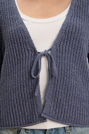 A close-up of a person wearing the THALIA CARDIGAN by Velvet by Graham & Spencer—a textured blue tie front design with rolled edges—layered over a white ribbed top and blue jeans.