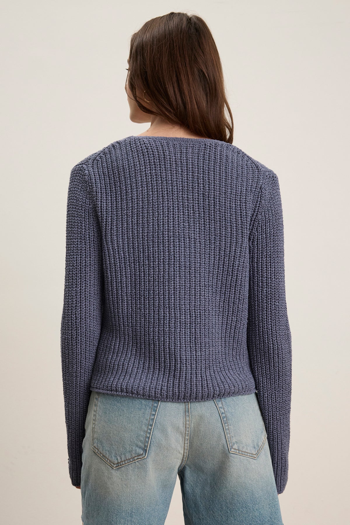 A person with long brown hair is seen from the back, wearing the THALIA CARDIGAN by Velvet by Graham & Spencer—a ribbed, tie-front purple cropped cardigan—paired with light blue jeans against a plain background.