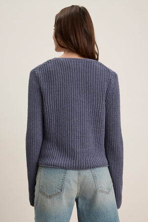A person with long brown hair is seen from the back, wearing the THALIA CARDIGAN by Velvet by Graham & Spencer—a ribbed, tie-front purple cropped cardigan—paired with light blue jeans against a plain background.