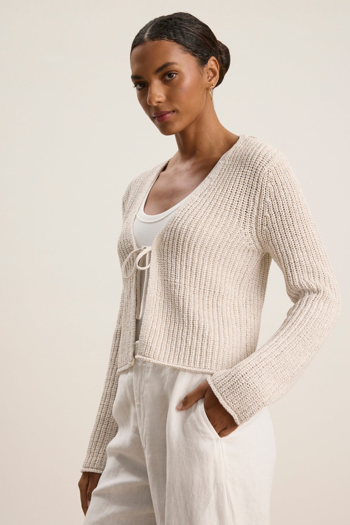 A woman wears the THALIA CARDIGAN by Velvet by Graham & Spencer—a light beige cropped style with rolled edges—over a white tank top and high-waisted white pants, standing with one hand in her pocket against a plain background.