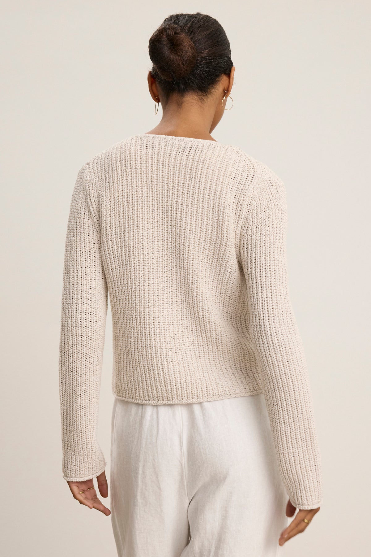 A person with hair in a bun, wearing the THALIA CARDIGAN by Velvet by Graham & Spencer and white pants, stands facing away from the camera against a plain background.-39111854096577