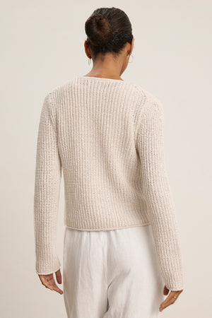 A person with hair in a bun, wearing the THALIA CARDIGAN by Velvet by Graham & Spencer and white pants, stands facing away from the camera against a plain background.