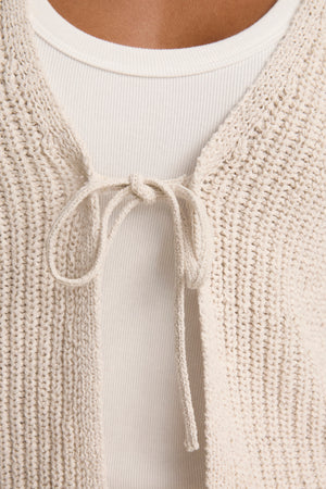Close-up of the THALIA CARDIGAN by Velvet by Graham & Spencer—textured beige, cropped with rolled edges, tied at the front in a bow and styled over a ribbed white top.