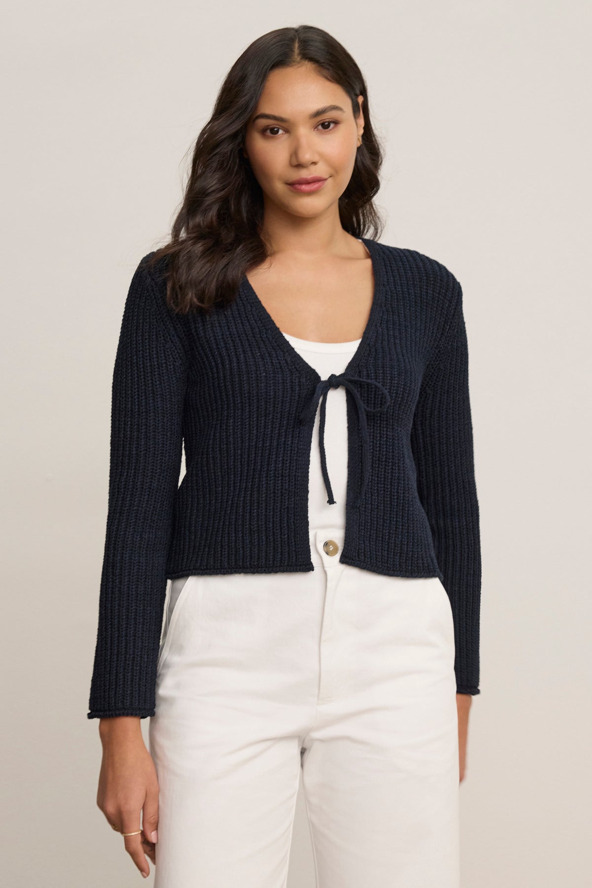 A woman with long dark hair wears the Velvet by Graham & Spencer THALIA CARDIGAN in navy blue over a white tank top and white pants, standing against a plain light background.