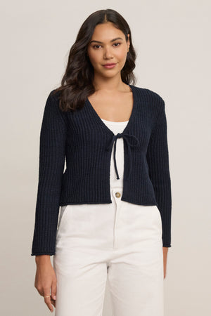 A woman with long dark hair wears the Velvet by Graham & Spencer THALIA CARDIGAN in navy blue over a white tank top and white pants, standing against a plain light background.