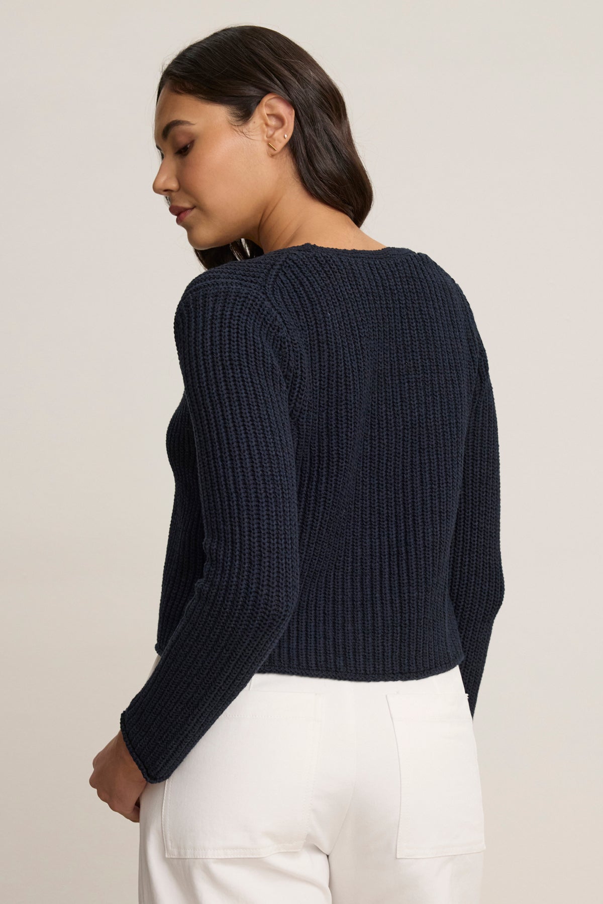 A woman stands against a neutral background, facing slightly away, wearing the THALIA CARDIGAN by Velvet by Graham & Spencer in navy blue ribbed fabric with a tie front, paired with white pants.-39120167502017