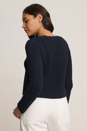 A woman stands against a neutral background, facing slightly away, wearing the THALIA CARDIGAN by Velvet by Graham & Spencer in navy blue ribbed fabric with a tie front, paired with white pants.