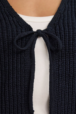 Close-up of a person wearing the Velvet by Graham & Spencer THALIA CARDIGAN, a navy blue piece with a stylish tie front fastened in a neat bow, layered over a white ribbed top.