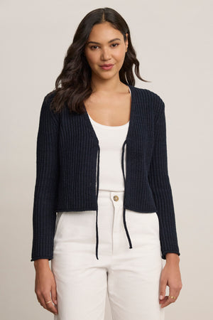 A woman faces forward, wearing the THALIA CARDIGAN by Velvet by Graham & Spencer in navy blue over a white top and white pants against a plain background.