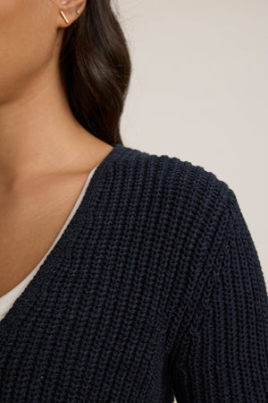 A close-up of a person wearing the THALIA CARDIGAN by Velvet by Graham & Spencer in dark blue over a white top, with part of their face and a gold ear cuff also visible.