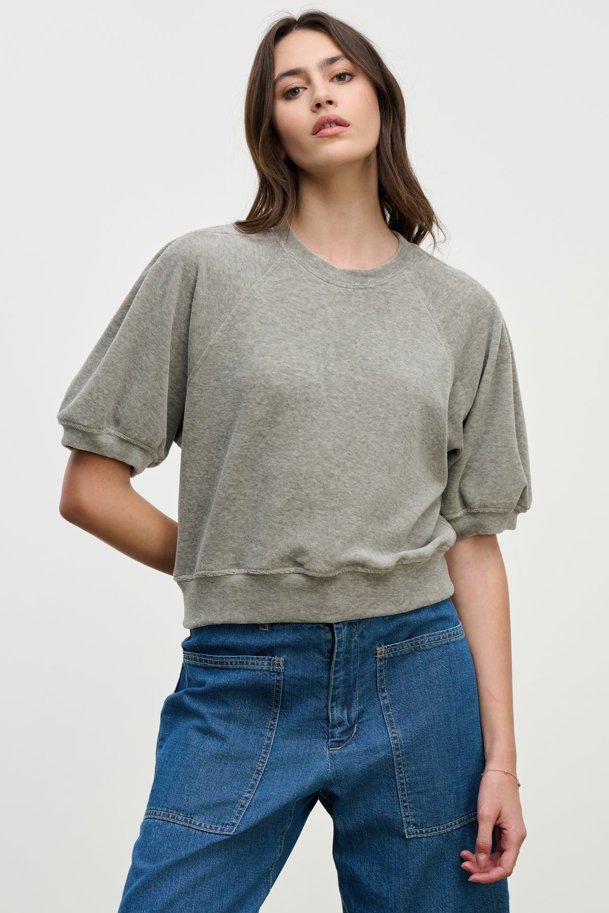 A woman wears the AVIS PULLOVER by Velvet by Graham & Spencer, a loose-fitting gray cotton velour top with raglan puff sleeves and a crew neckline, paired with blue jeans against a plain light background.-39814820790465
