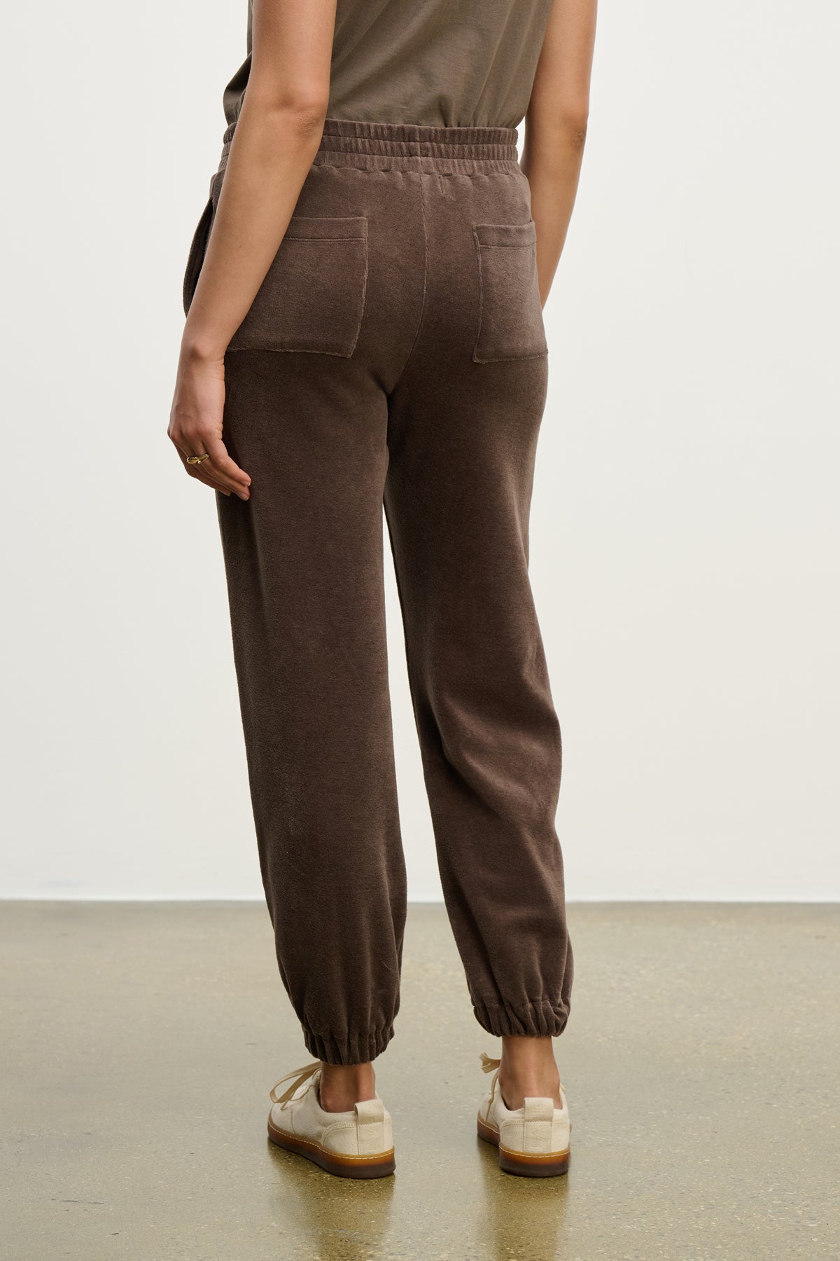 A person stands on a smooth floor, facing away from the camera, wearing Velvet by Graham & Spencer's ESSIE SWEATPANT with back and side seam pockets, paired with beige sneakers.-39814830489793