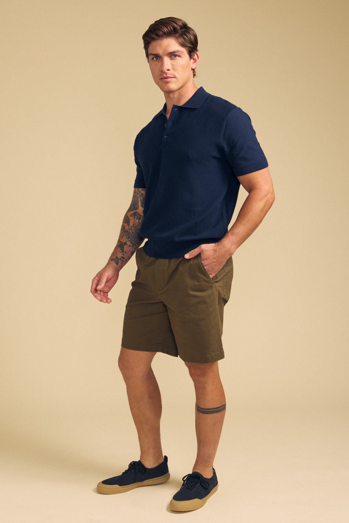 A man stands against a beige background wearing Velvet by Graham & Spencer's GARTH SHORT, a navy short-sleeve polo, navy sneakers, and showing visible arm and leg tattoos.-41701930893505