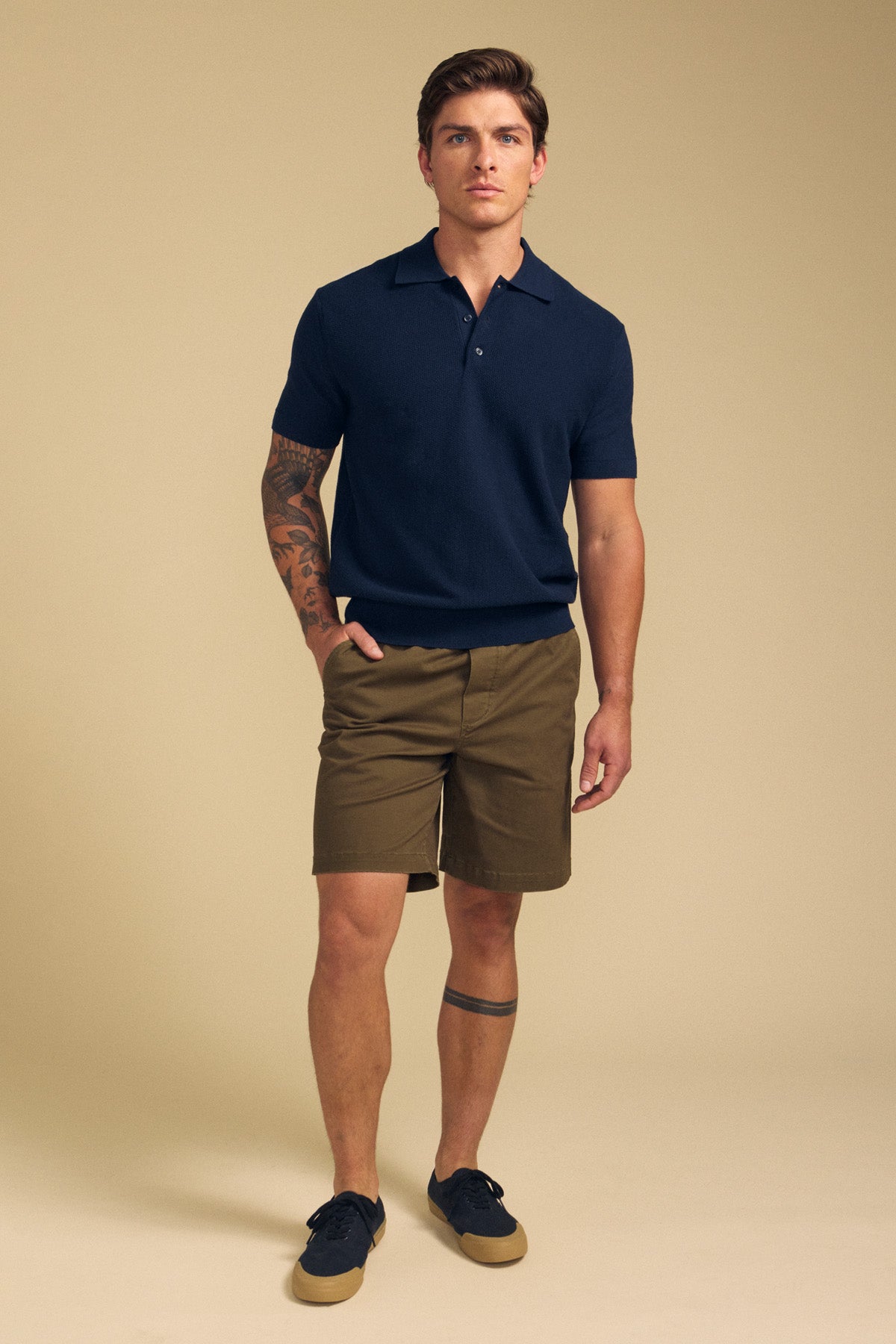 A man stands against a beige background wearing the DILLON SWEATER POLO by Velvet by Graham & Spencer, styled with olive shorts and navy sneakers. Tattoos on his right arm and left leg complete this effortless warm-weather look.-41701930533057