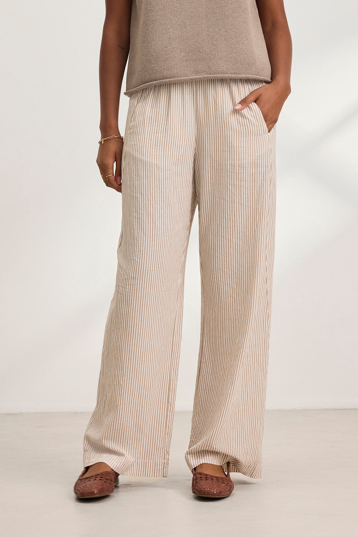 A person wearing the KELLY PANT by Velvet by Graham & Spencer—beige and white striped, elastic waist, wide-leg—paired with a taupe sleeveless top, brown woven shoes, and a bracelet stands indoors with one hand in their pocket.