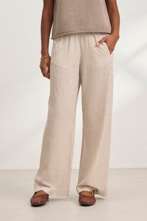 A person wearing the KELLY PANT by Velvet by Graham & Spencer—beige and white striped, elastic waist, wide-leg—paired with a taupe sleeveless top, brown woven shoes, and a bracelet stands indoors with one hand in their pocket.