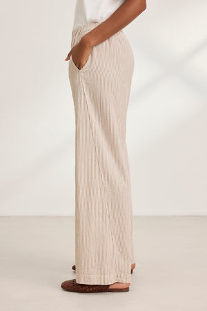 Person in Velvet by Graham & Spencer's KELLY PANT (beige and white wide-leg), hands in pockets, paired with brown woven sandals. Side view against a plain background.