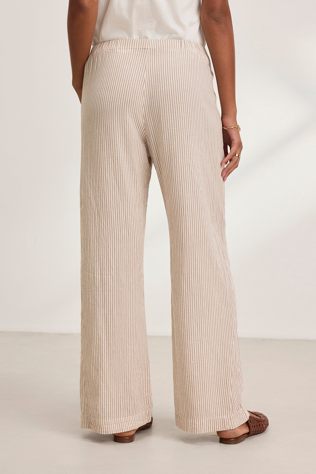 A person is seen from behind wearing the KELLY PANT by Velvet by Graham & Spencer, featuring beige and white stripes and wide legs, paired with brown shoes on a white floor.