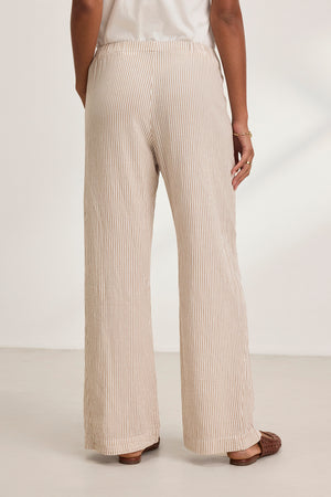 A person is seen from behind wearing the KELLY PANT by Velvet by Graham & Spencer, featuring beige and white stripes and wide legs, paired with brown shoes on a white floor.