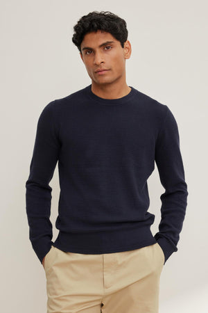 A person wearing the ACE COTTON CASHMERE THERMAL CREW by Velvet by Graham & Spencer and beige pants stands against a plain background, hands in pockets.