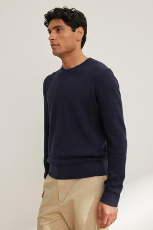 A person in the ACE COTTON CASHMERE THERMAL CREW by Velvet by Graham & Spencer and beige pants stands against a plain background, exuding timeless elegance.